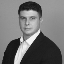 Headshot of a man in a black suit and white shirt with short dark hair on a gray background.