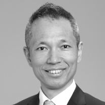 Smiling middle-aged man wearing suit and tie against a plain gray background.