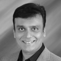 Smiling man with short dark hair wearing a checkered blazer and dark shirt on a gray background.