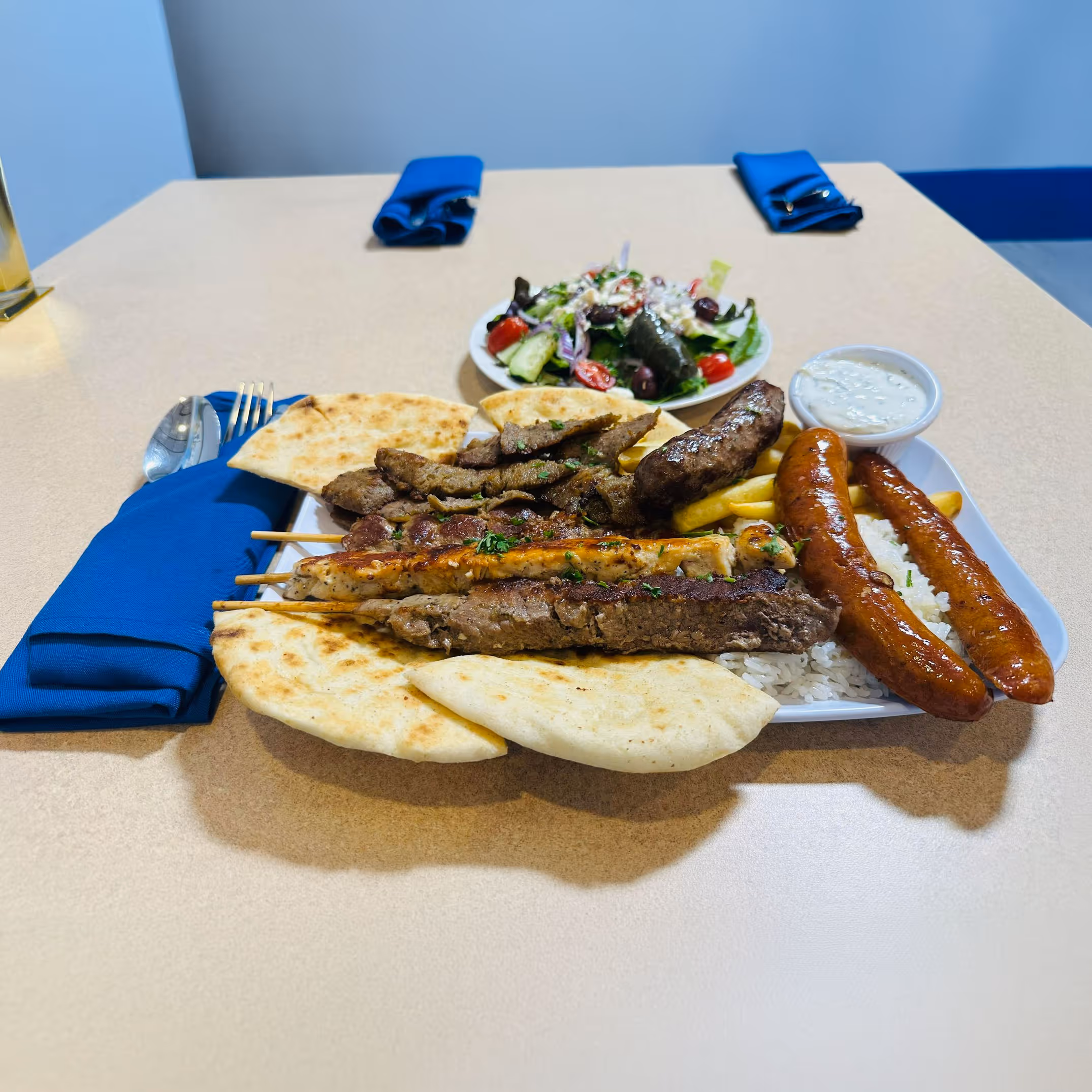 Plate of Greek mixed grill platter with pita bread, grilled meat skewers, sausages, rice, fries, tzatziki sauce, and a side salad with olives and tomatoes.