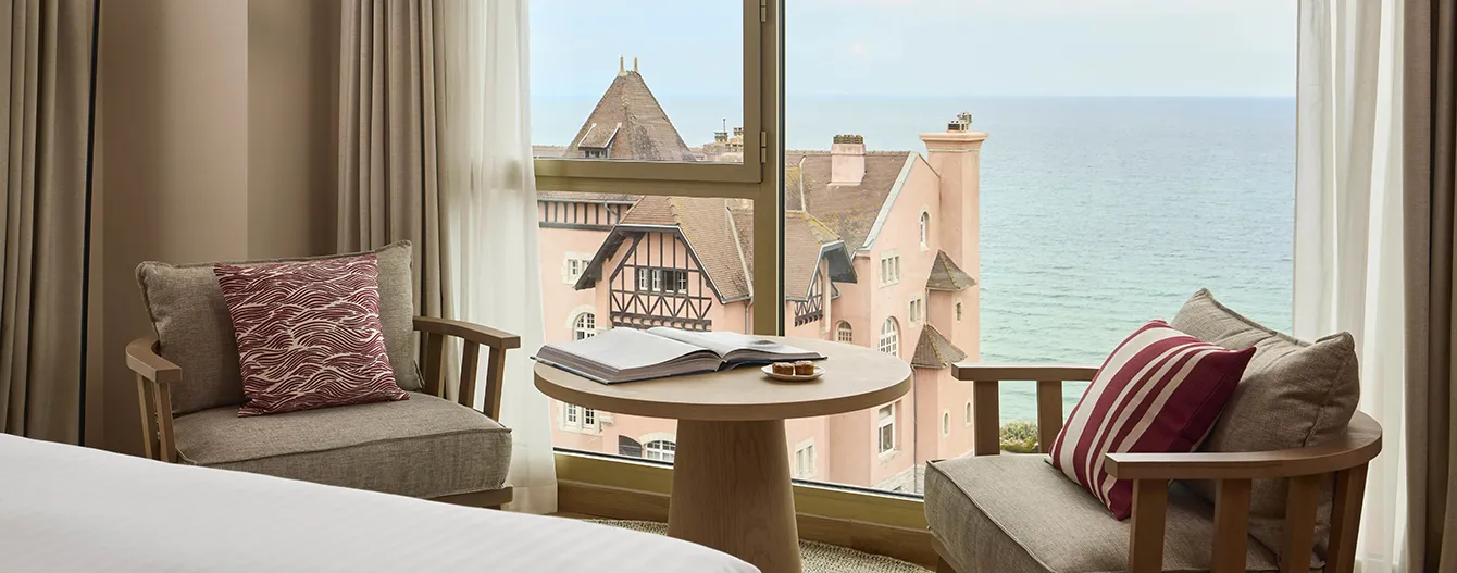 Vue depuis une chambre avec deux fauteuils, une table ronde, un livre ouvert, et la mer au loin.