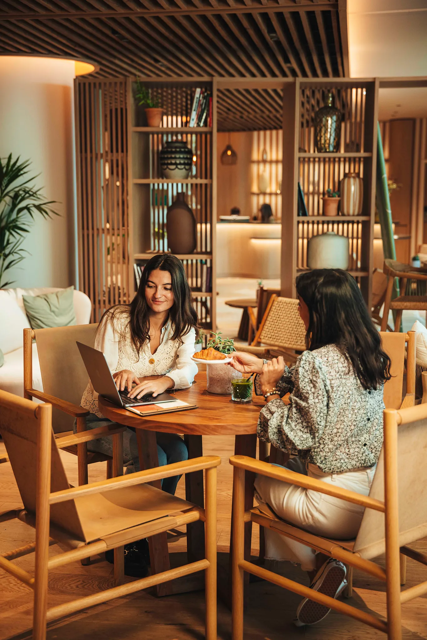 Deux femmes discutent autour d'une table en bois dans un café chaleureux et moderne.