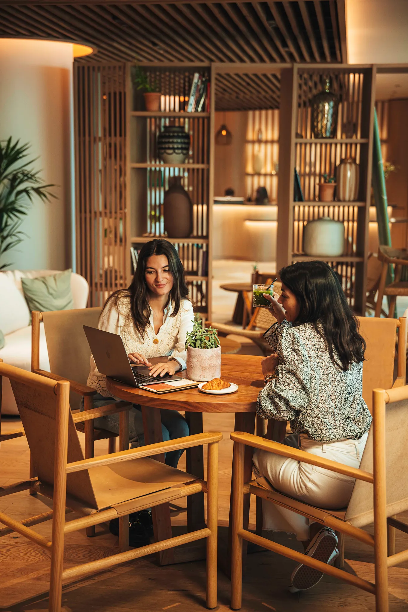Deux femmes discutant à une table en bois avec un ordinateur portable, plante et croissant à côté.