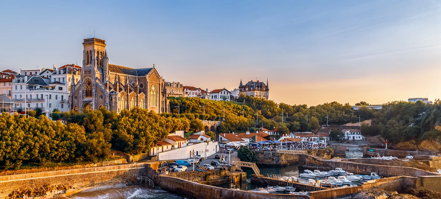 Vue côtière au coucher du soleil avec une grande église et un port de plaisance rempli de bateaux.