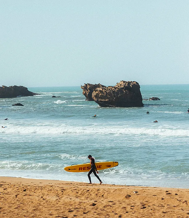 Un sauveteur marche sur la plage avec une planche de sauvetage jaune près des rochers en mer.