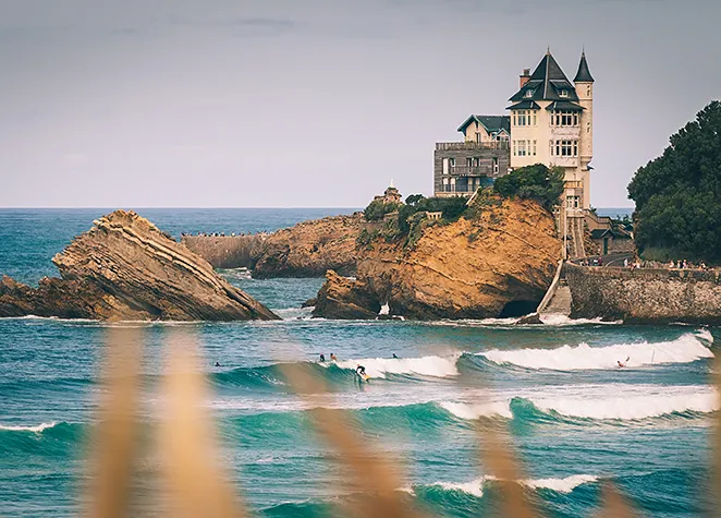 Maison sur une falaise rocheuse au bord de l'océan avec des surfeurs dans les vagues.