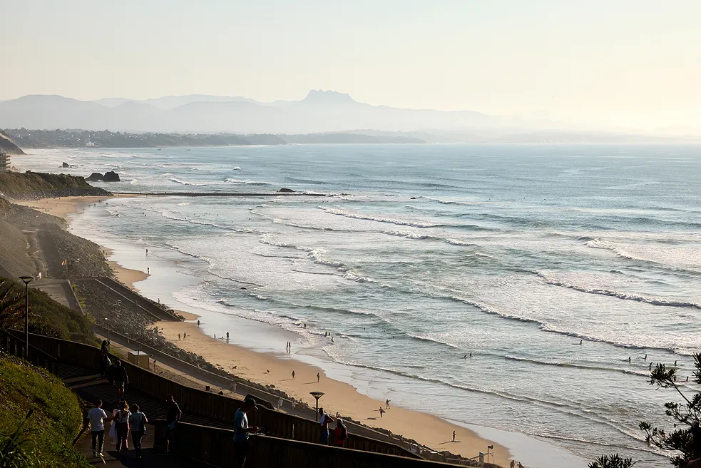 Plage avec vagues et personnes sur le sable au coucher du soleil, avec montagnes en arrière-plan.