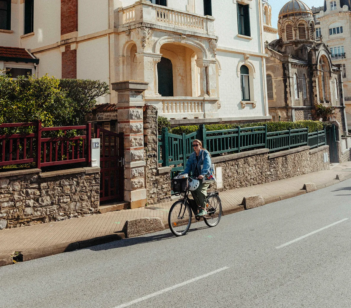 Une femme portant une veste en jean roule à vélo sur une rue devant des maisons anciennes.