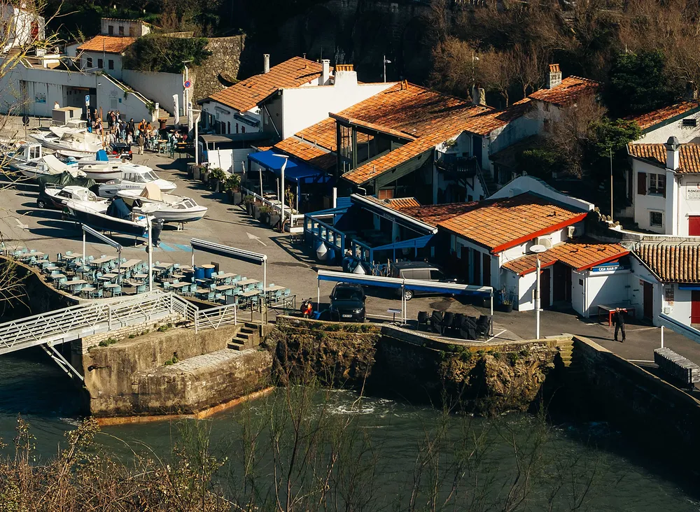 Petit port avec bateaux, tables de terrasse et bâtiments à toits rouges au bord de l'eau.