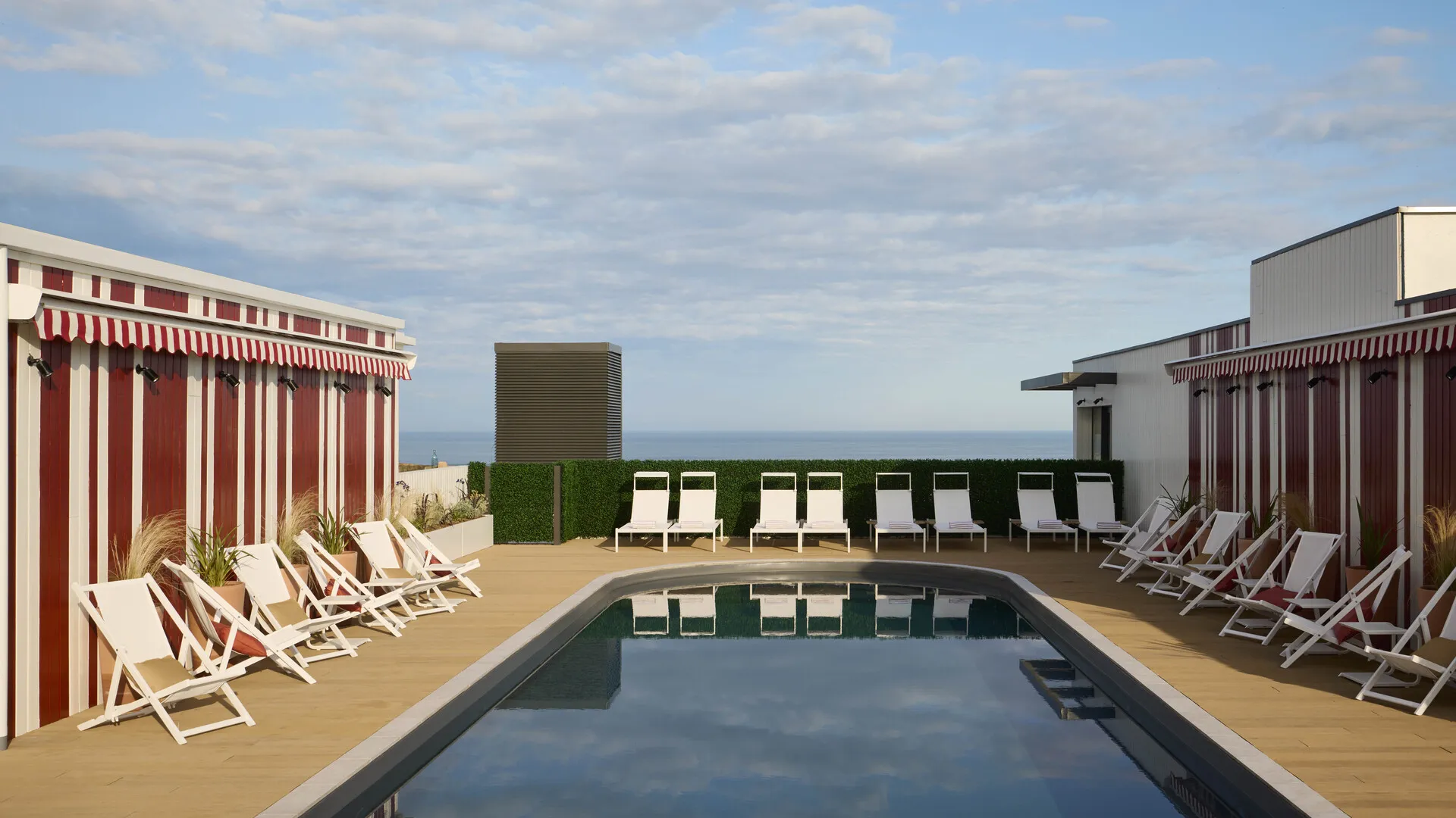 Piscine bordée de chaises longues blanches et de cabines rayées rouges et blanches, vue sur l'océan.
