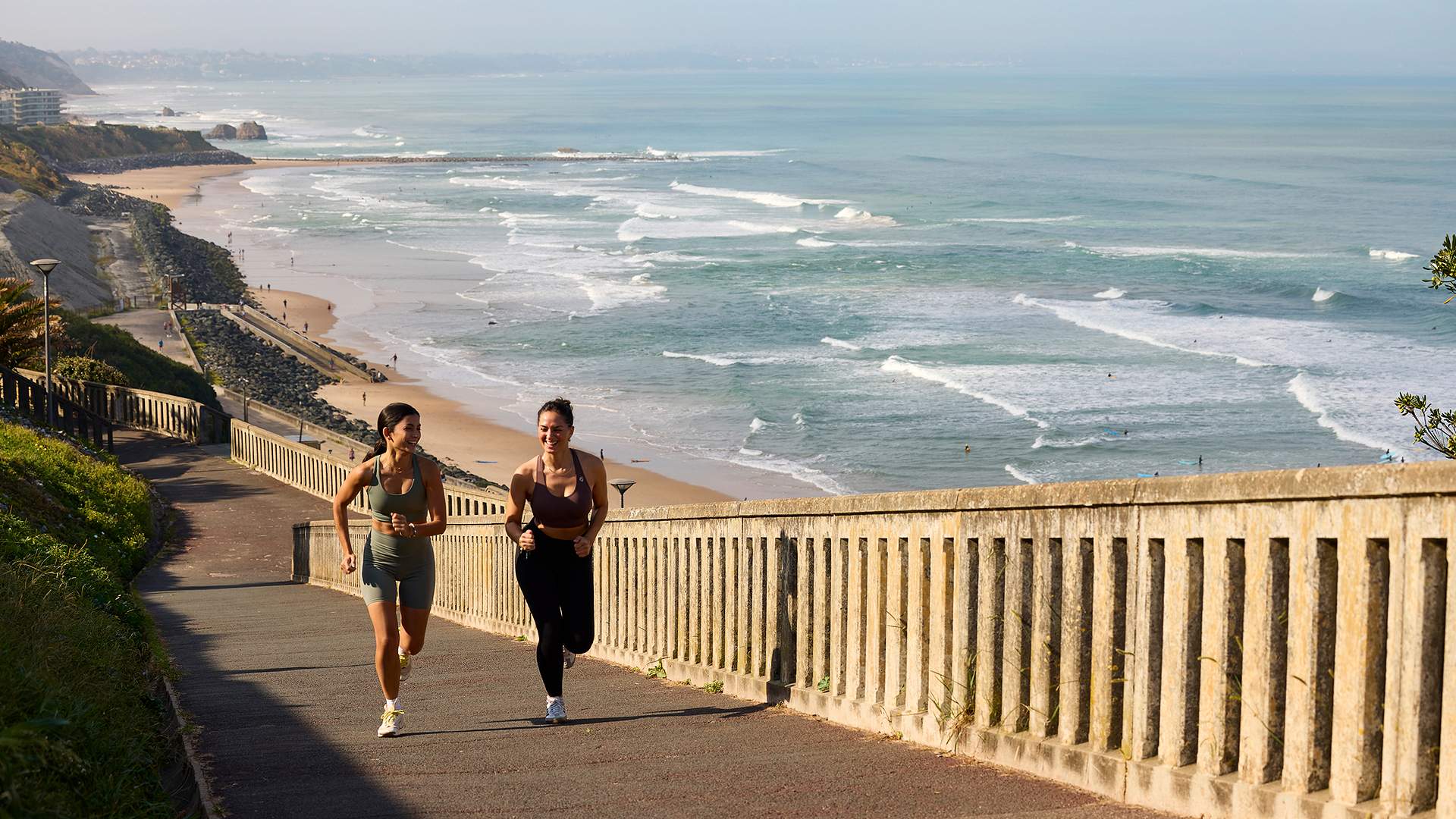 Deux femmes courent sur un sentier en bord de mer avec plage et vagues à l'arrière-plan.