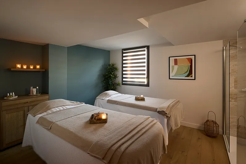 Salle de massage avec deux tables, bougies allumées, plante verte et tableau abstrait au mur.