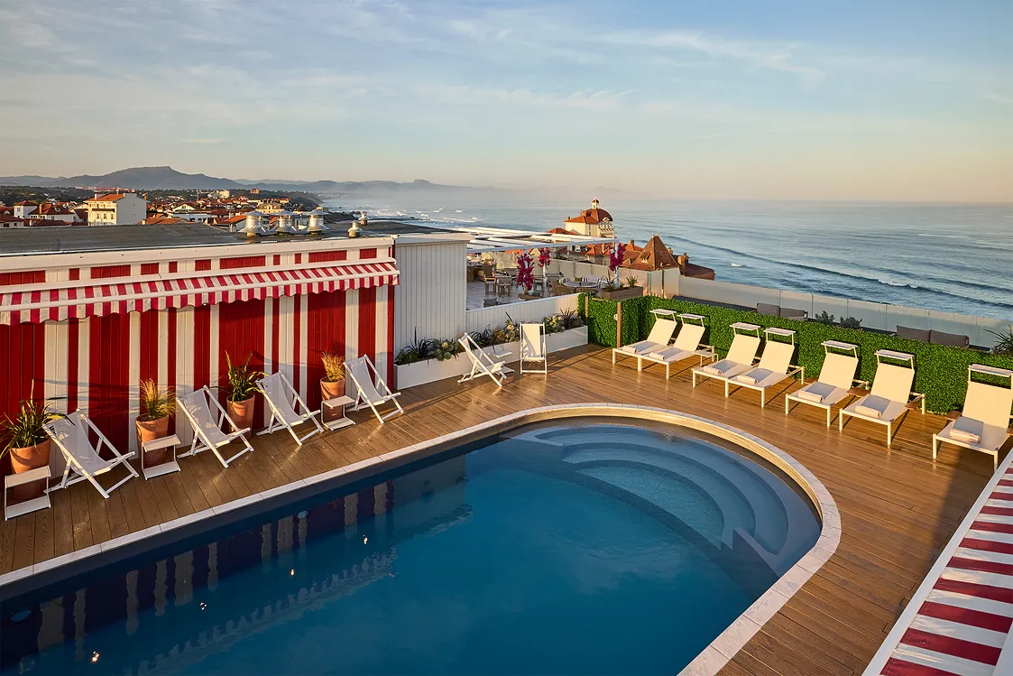 Piscine sur un toit avec chaises longues blanches et vue sur l'océan au coucher du soleil.