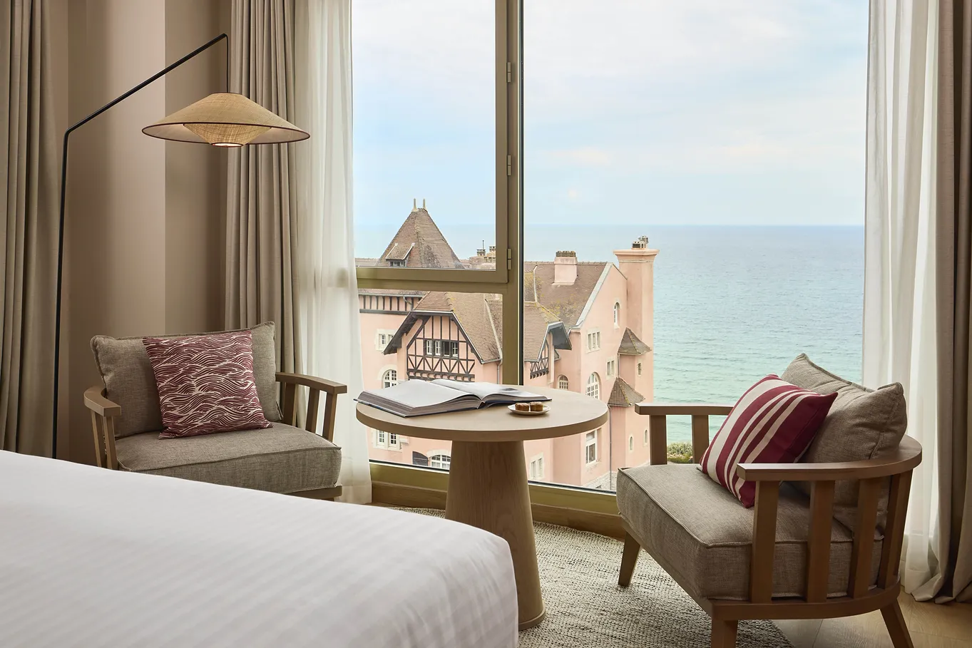 Coin salon avec chaises, table ronde et vue sur maison au bord de la mer à travers une fenêtre.