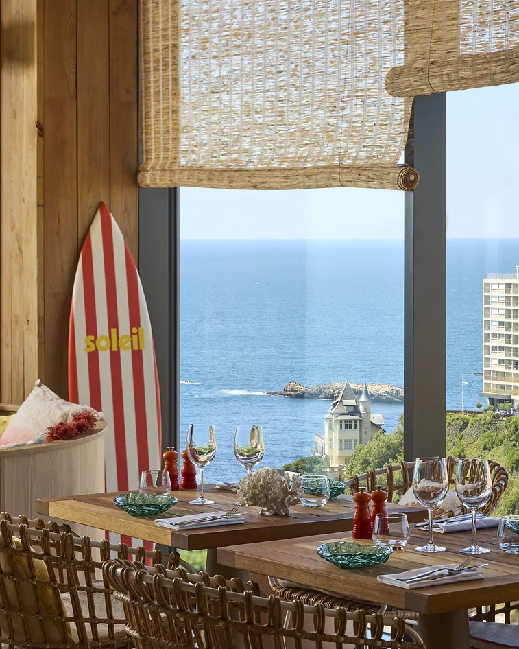 Dining room overlooking the ocean, red striped surfboard, set wooden tables.