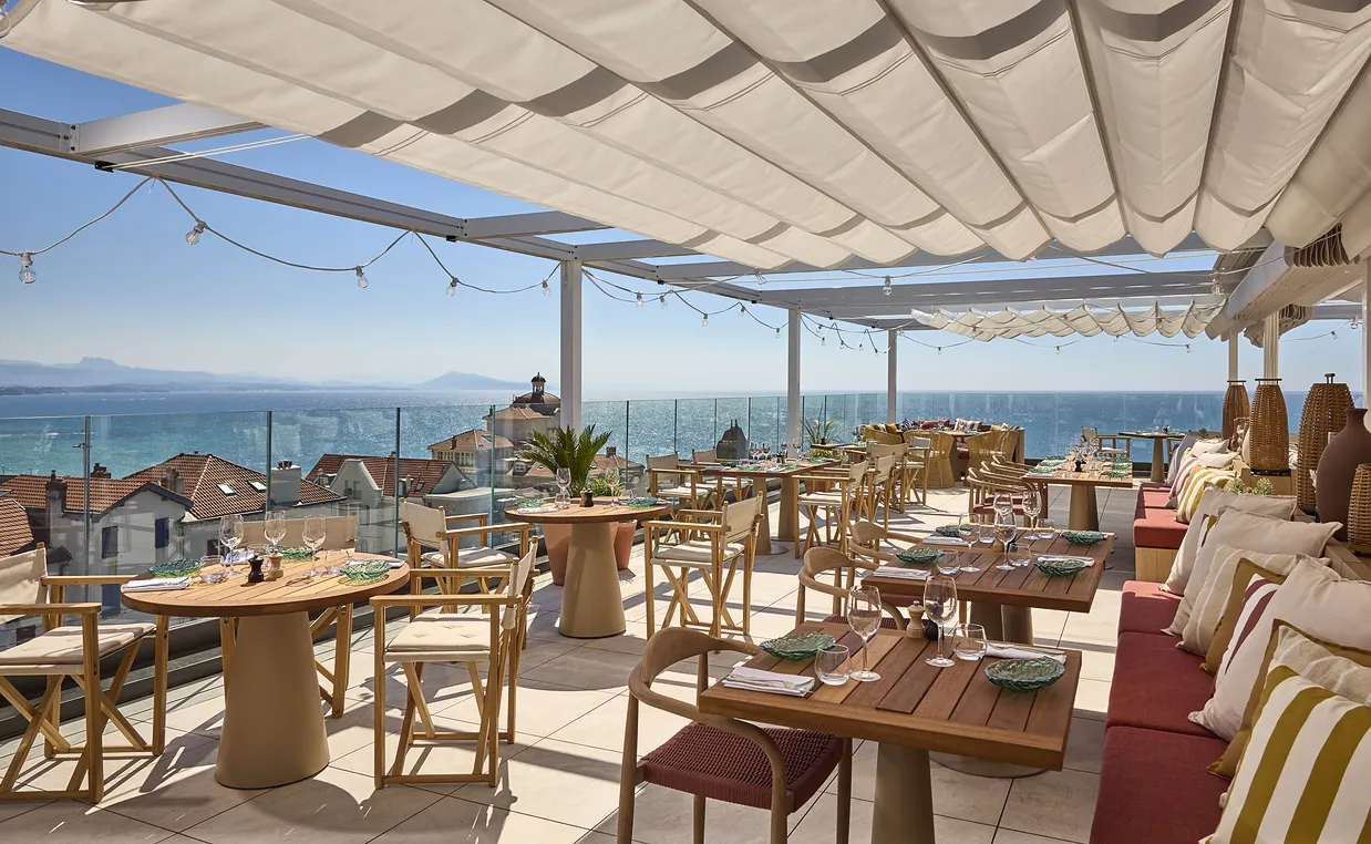 Terrasse de restaurant élégante avec tables en bois et vue sur mer sous un toit ombragé.