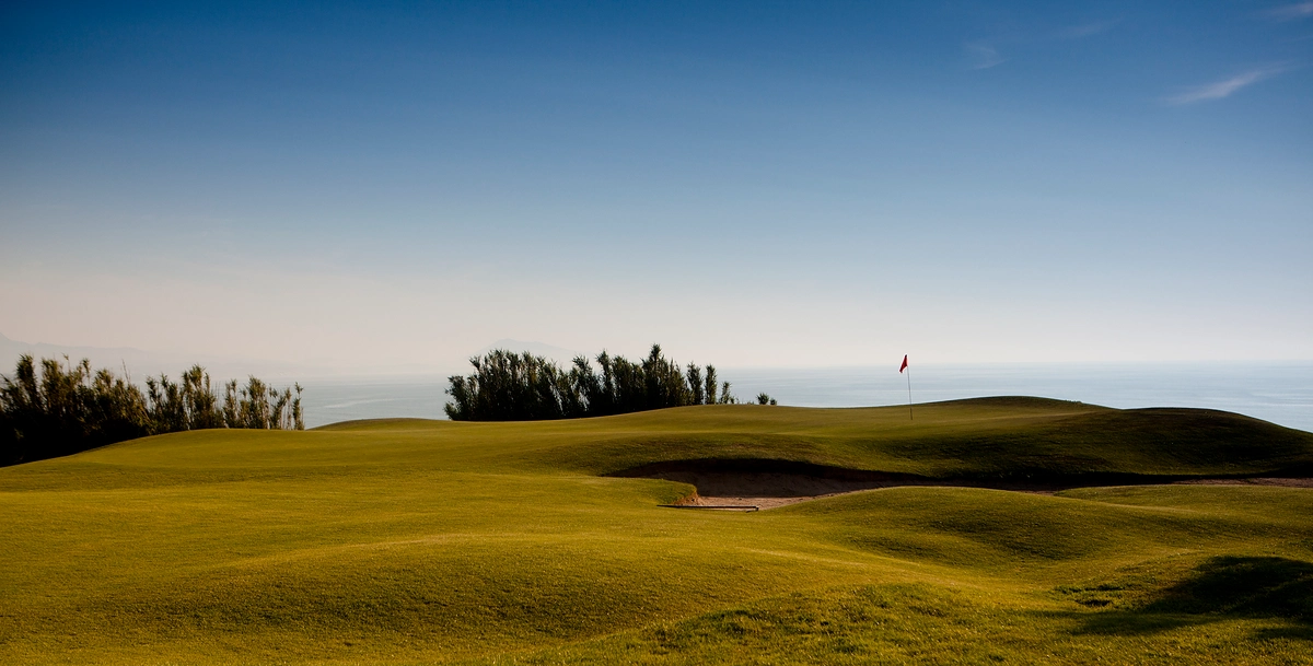 Hilly golf course with a red flag, bushes in the background and ocean views.