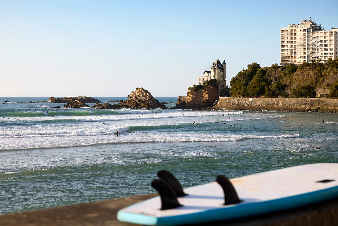 Planche de surf posée avec vue sur des surfeurs montant les vagues près de rochers et bâtiments en bord de mer.