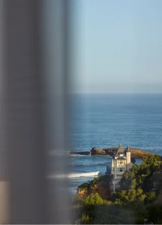 Vue sur une maison sur une colline bordant la mer avec une fenêtre floue au premier plan.