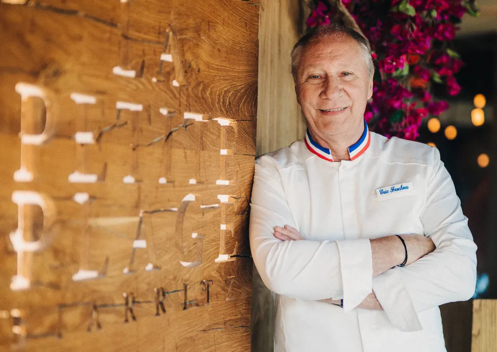 Chef souriant en veste blanche avec col tricolore, bras croisés, devant un mur en bois gravé.