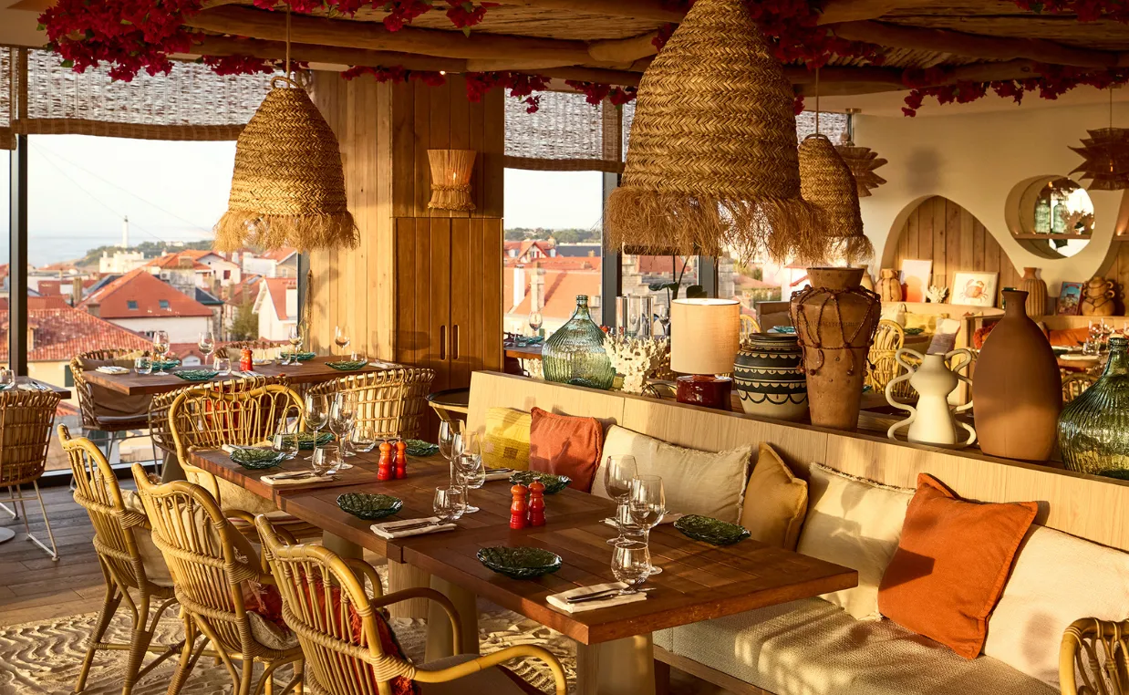 Restaurant chaleureux avec tables en bois, chaises en osier, lampes suspendues en paille et coussins colorés.