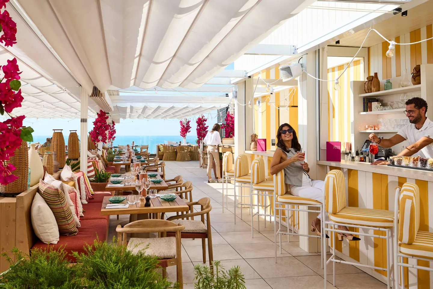 Terrasse de bar avec chaises jaunes et blanches, femme buvant un cocktail et serveurs en arrière-plan.
