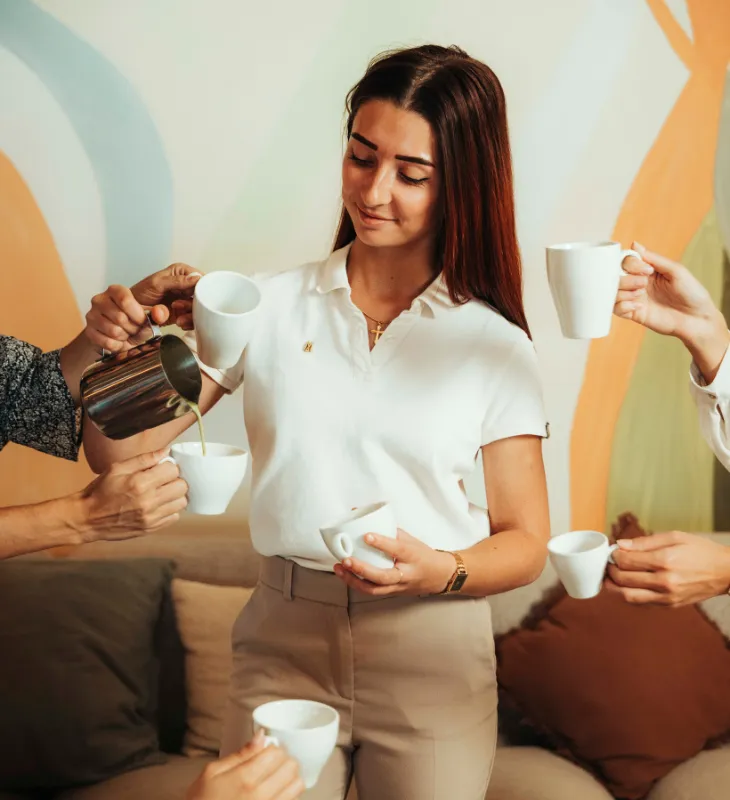 Une femme verse du café dans une tasse tenue par une autre personne, entourée de plusieurs mains tenant des tasses blanches.