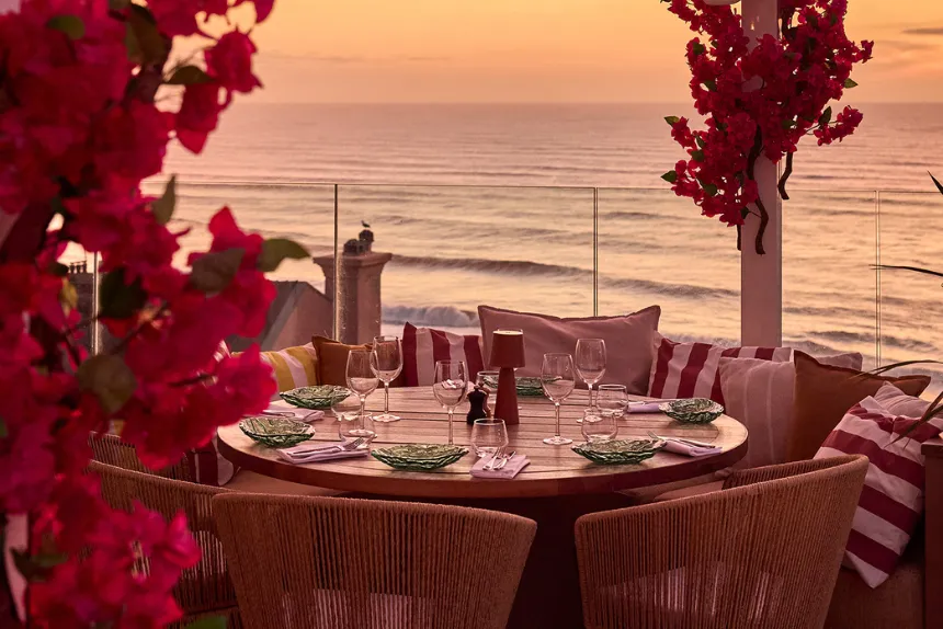 Round table set with dishes overlooking the ocean at sunset decorated with red flowers.