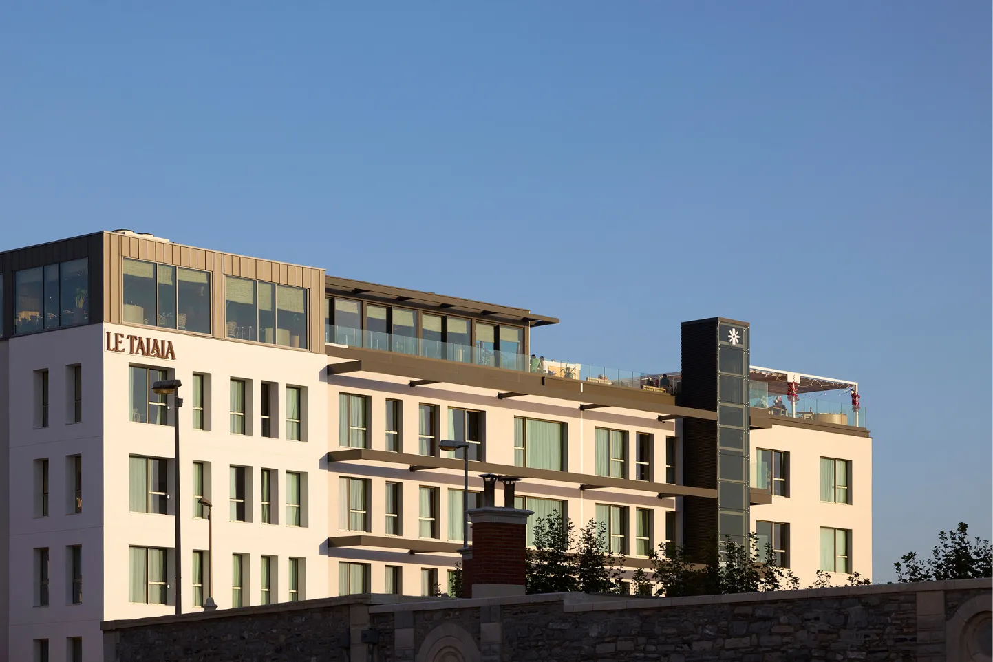 Bâtiment moderne d'hôtel Le Talaia avec terrasse vitrée sous ciel bleu dégagé.