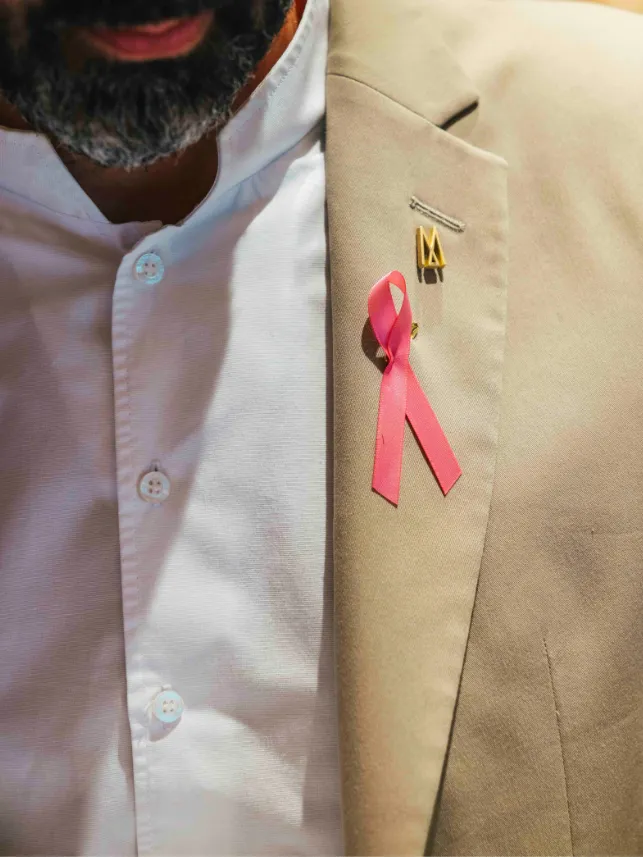 Un homme en chemise blanche et veste beige porte un ruban rose symbolisant la lutte contre le cancer du sein.