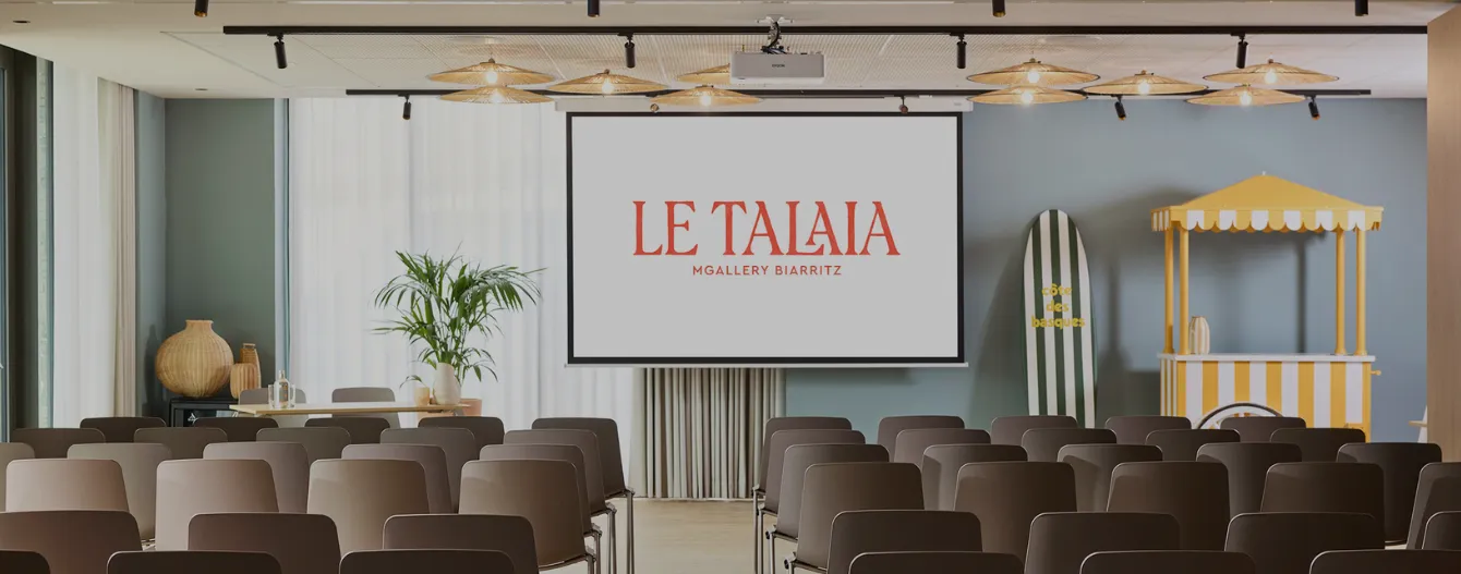 Salle de conférence avec chaises, écran affichant Le Talia, plante, surf et stand jaune rayé.