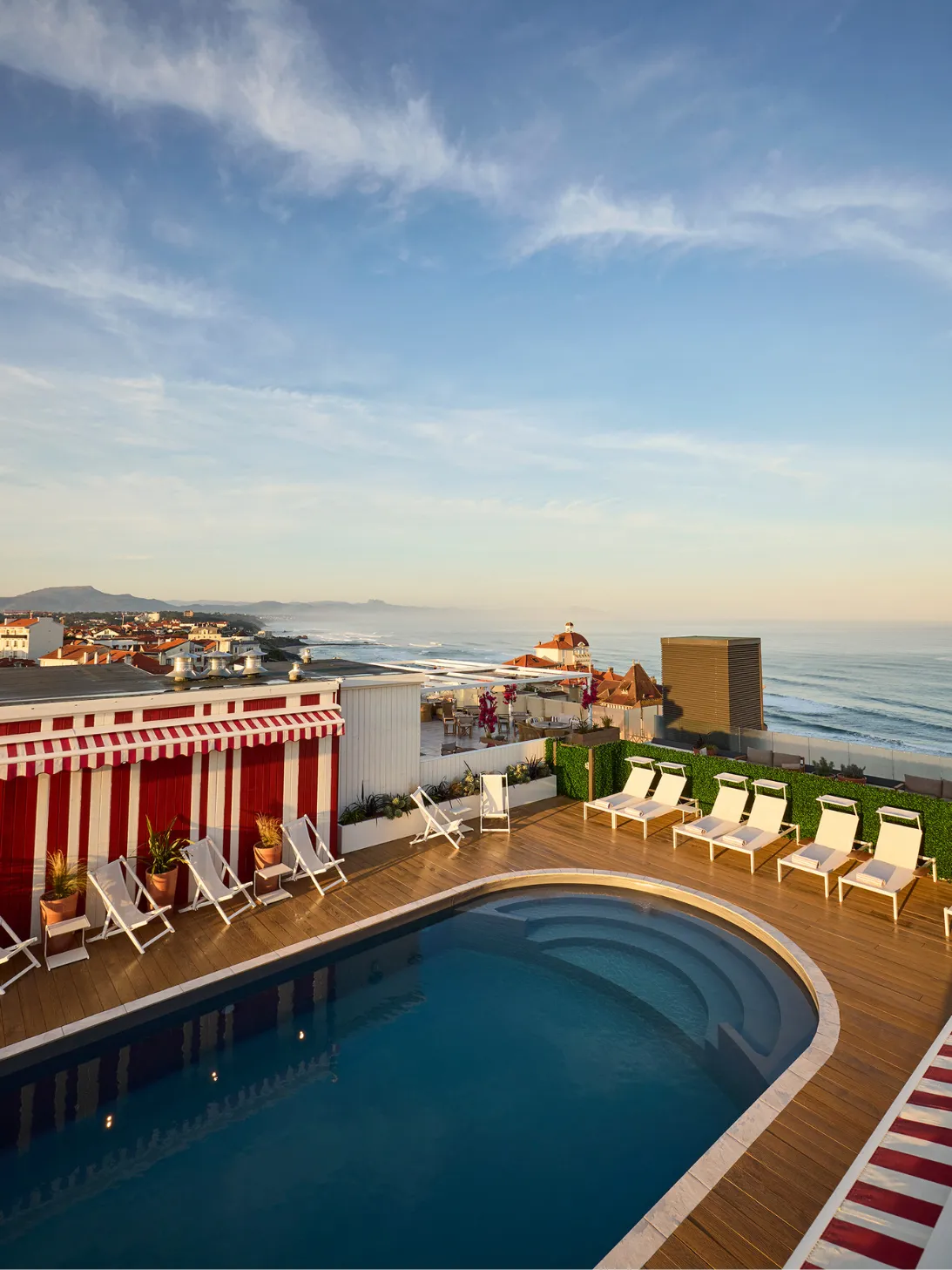 Piscine sur une terrasse avec chaises longues blanches, vue sur l'océan et le ciel au coucher du soleil.
