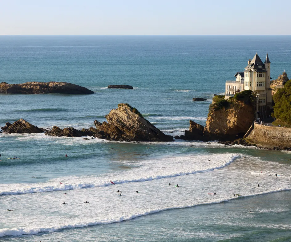 Plage avec des surfeurs dans l'eau et une maison sur une falaise rocheuse au bord de l'océan.