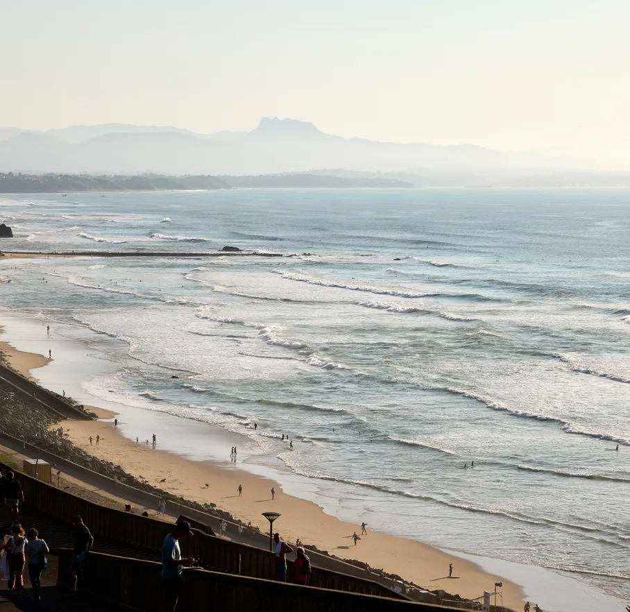 Plage avec vaguelettes, silhouettes de personnes sur le sable et montagnes à l'horizon.