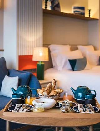 A hotel room breakfast tray featuring blue teapots, a basket of pastries and a baguette, orange juice, and jams, set on a coffee table in front of a bed with crisp white sheets, creating a cozy and warm ambiance.