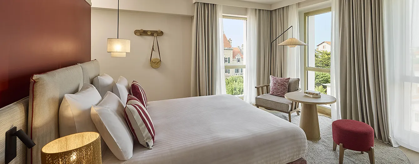 An elegant hotel room with a king-size bed, burgundy striped cushions, terracotta walls and a view over the rooftops of Biarritz
