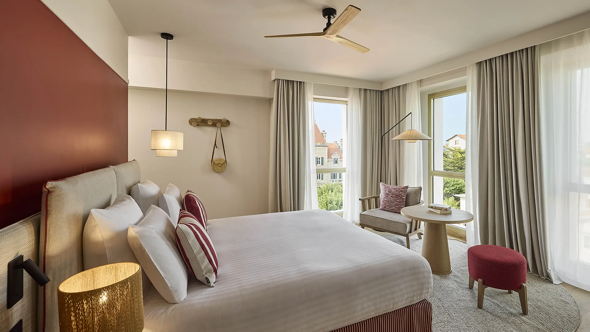Chambre d'hôtel avec grand lit, ventilateur au plafond, mur terracotta et fauteuil rose près de la fenêtre