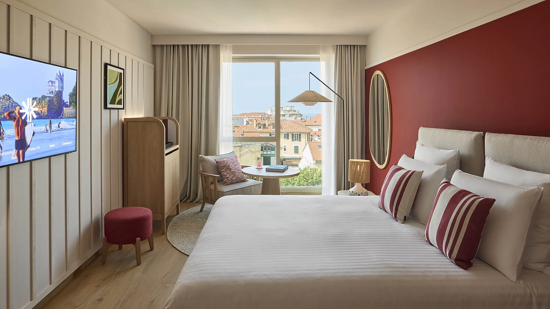 Chambre d'hôtel avec grand lit, télévision murale, mur bordeaux et vue sur la ville ensoleillée