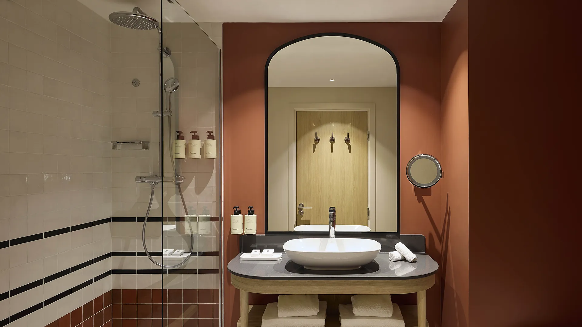 Salle de bain d'hôtel avec vasque, miroir cintré et carrelage terracotta, murs bordeaux