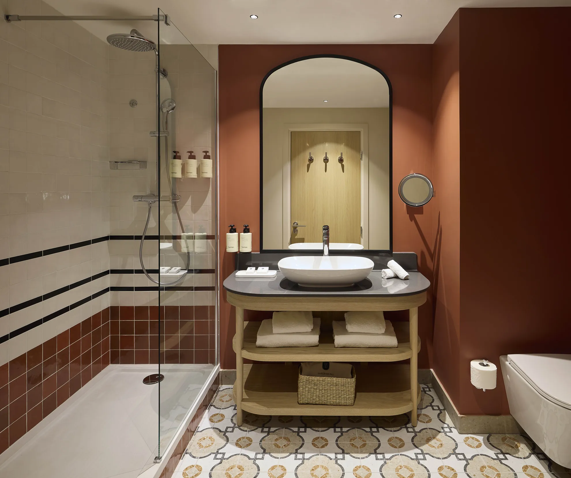 Salle de bain d'hôtel avec vasque sur meuble ouvert, douche vitrée et carrelage terracotta aux accents noirs