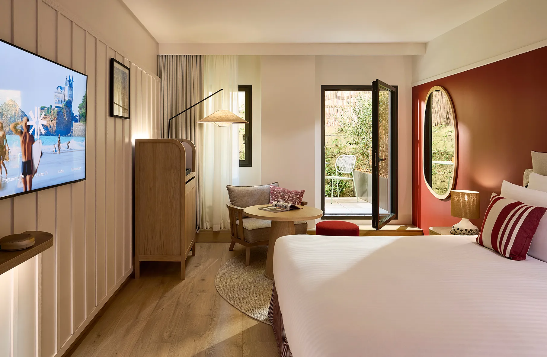 Chambre d'hôtel avec grand lit, télévision murale, mur terracotta et vue sur jardin