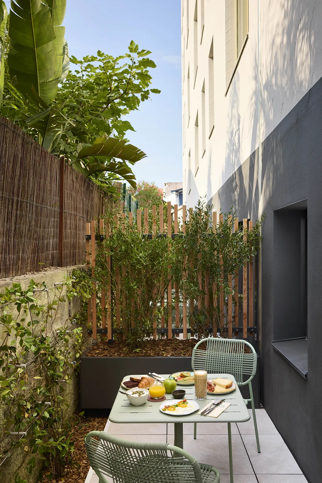 Terraza urbana de un hotel con una mesa puesta, sillas verdes y bambú entre dos fachadas