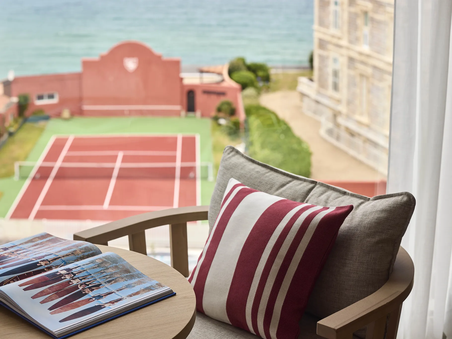 Terrasse d'hôtel avec coussin rayé rouge et blanc, vue sur un court de tennis et la mer en arrière-plan