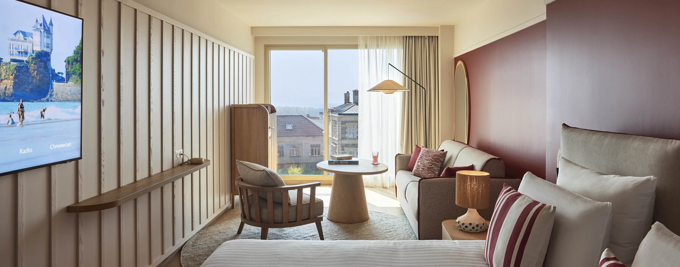 A room at the Le Talaia Mgallery Biarritz hotel with a bed, a seating area, a wall-mounted television and views of the town and the ocean