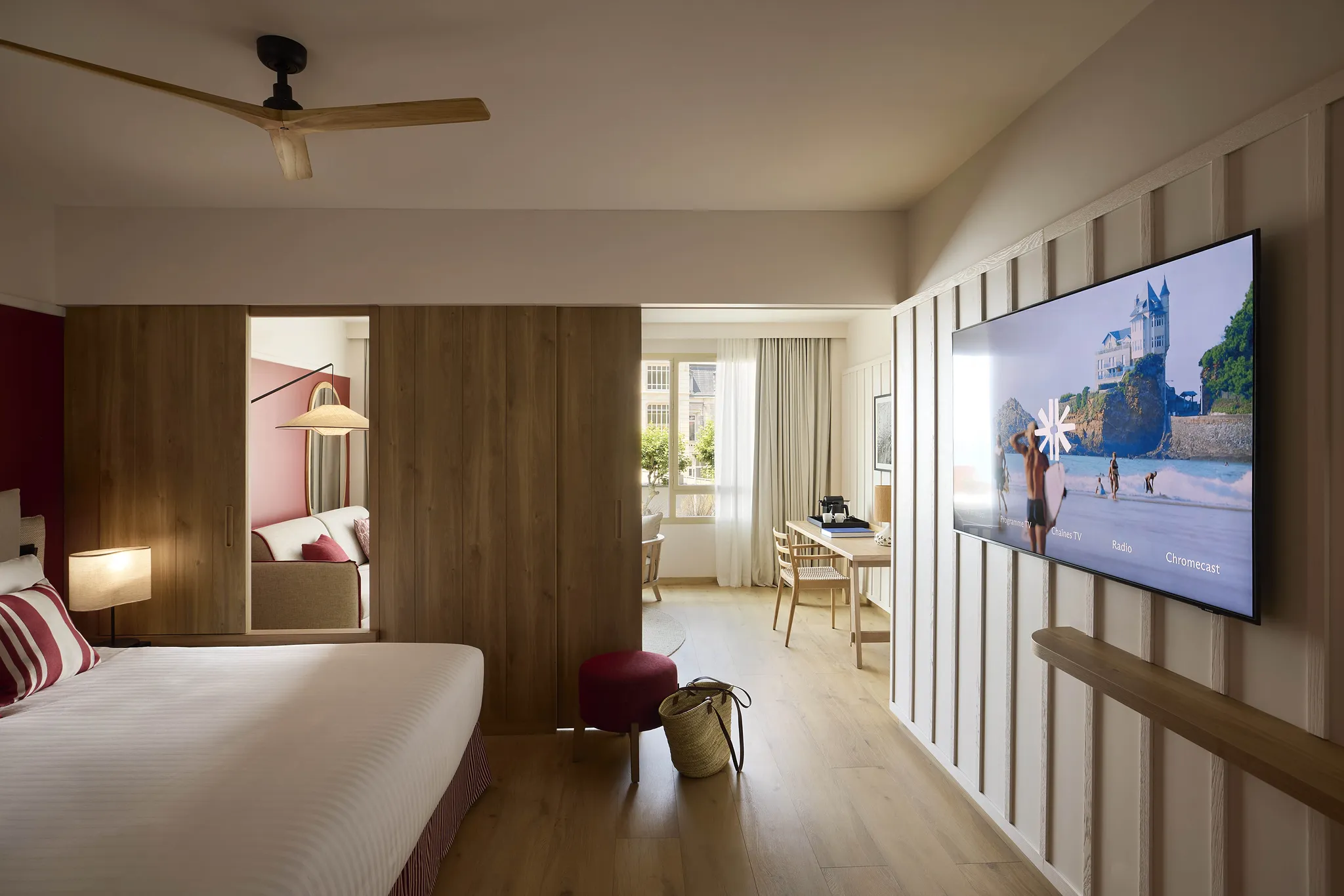 Vue d'ensemble d'une chambre de l'hôtel Le Talaia Mgallery Biarritz avec lit, télévision murale, cloisons en bois et ouverture sur le salon