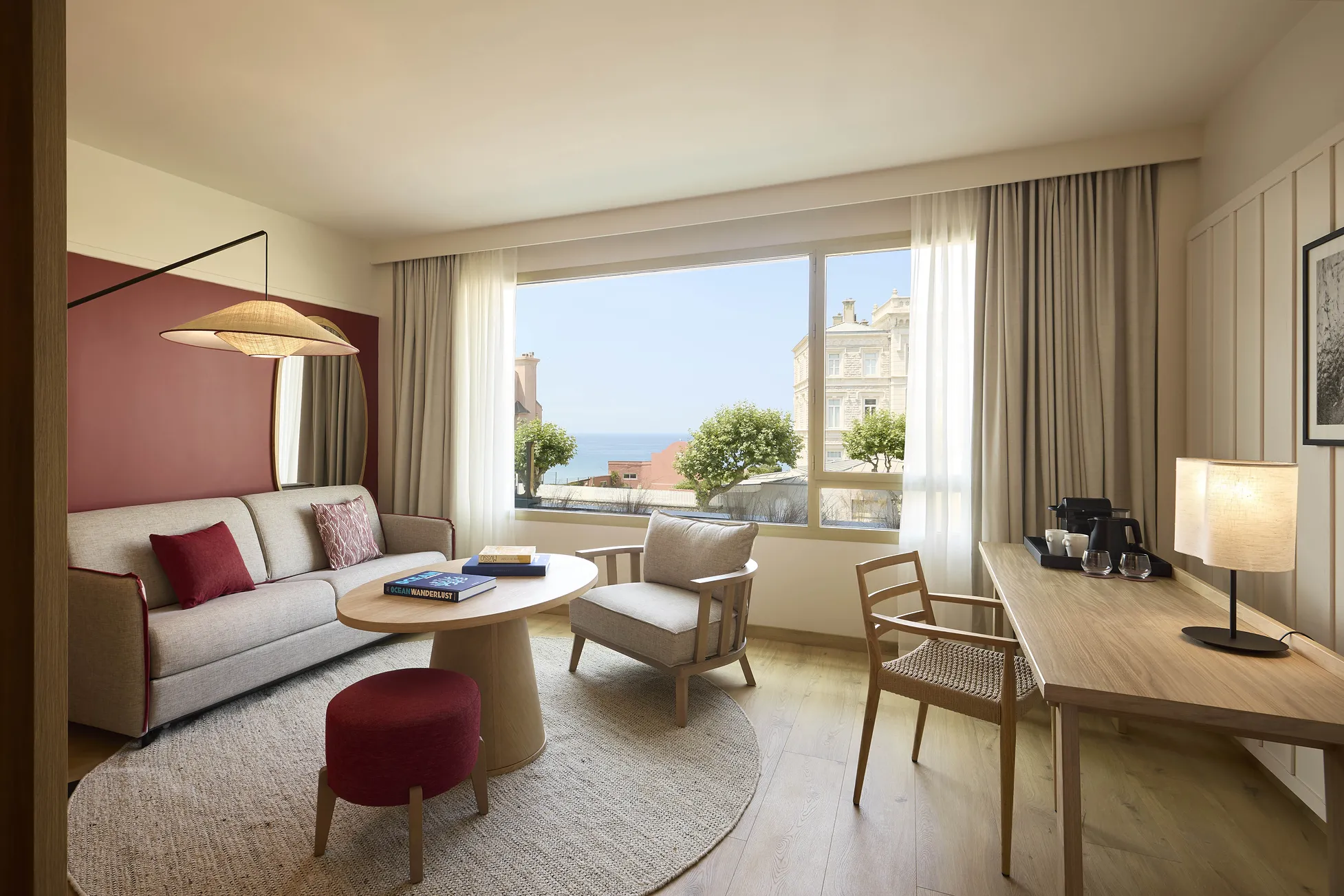 Suite lumineuse de l'hôtel Le Talaia Mgallery Biarritz avec canapé beige, accents bordeaux et vue panoramique sur l'océan
