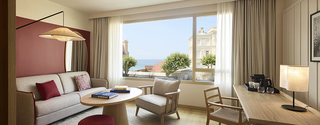 The living area of a one-bedroom room at the Le Talaia Mgallery hotel in Biarritz, featuring a beige sofa, burgundy cushions, a wooden coffee table and a view of the ocean through the large window