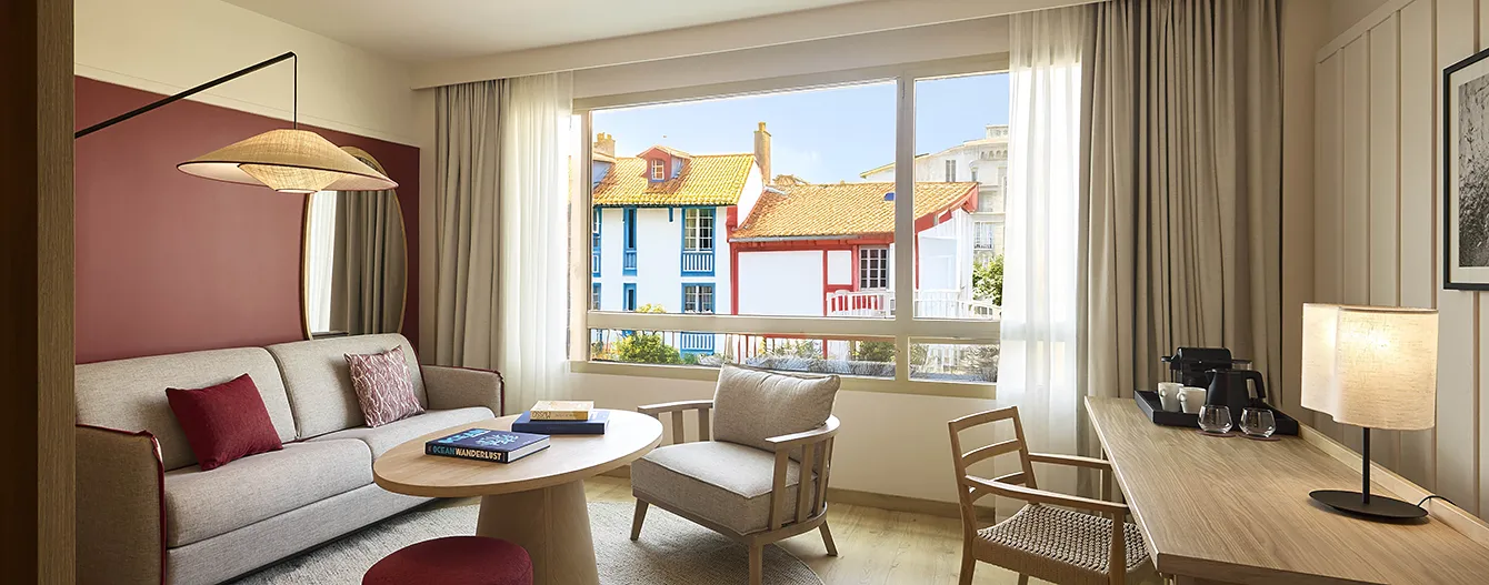 The living room of a suite at the Le Talaia Mgallery hotel in Biarritz, featuring a beige sofa, burgundy accents and a view of the colourful Basque houses