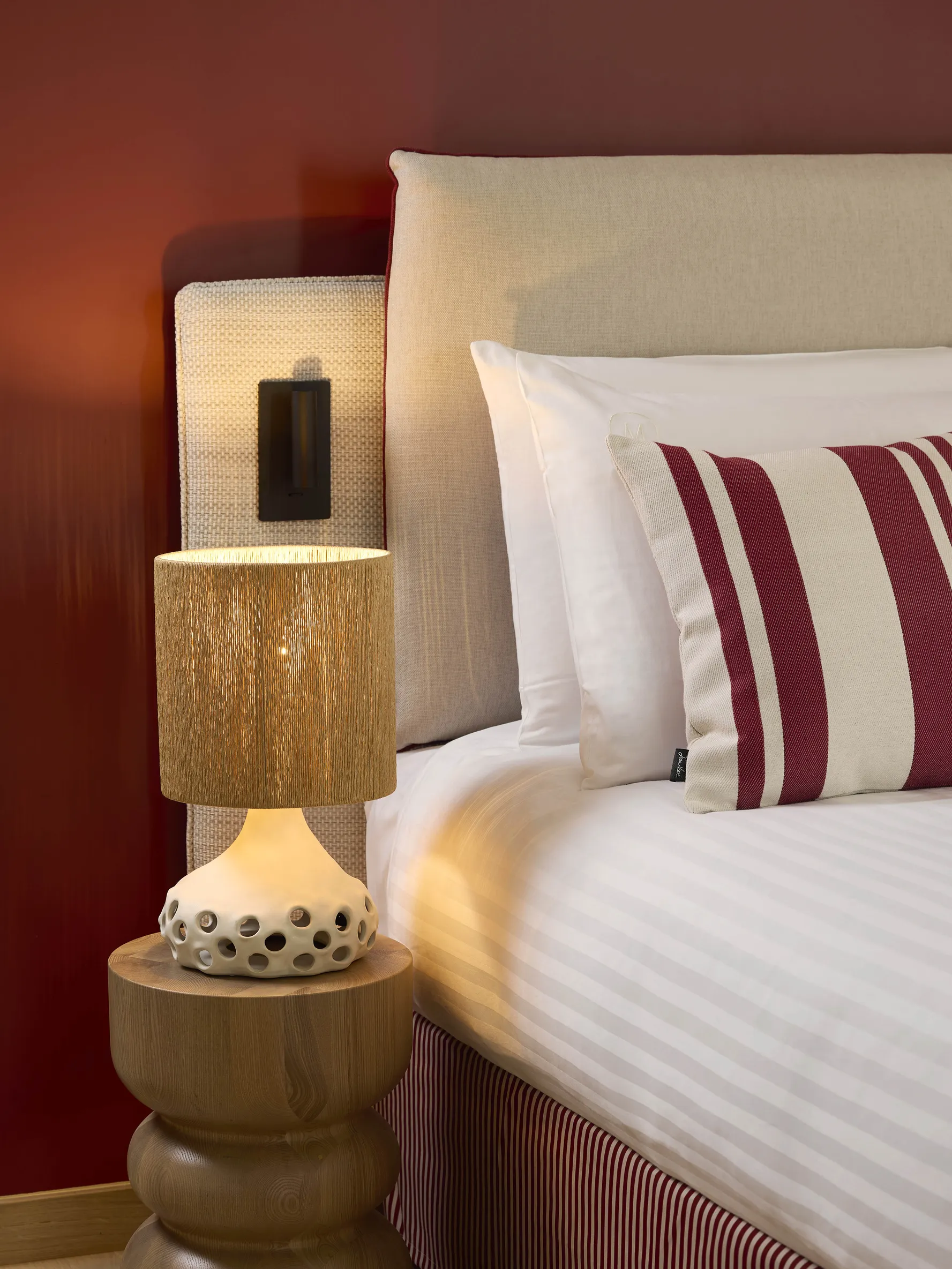 Détail du chevet d'une chambre de l'hôtel Le Talaia Mgallery Biarritz avec lampe en bois naturel et oreillers rayés bordeaux
