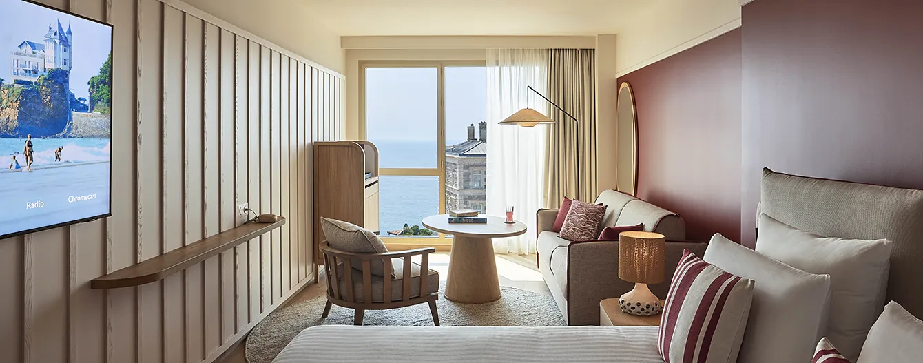 A room at the Le Talaia Mgallery Biarritz hotel with a seating area, a wall-mounted television and a view of the ocean through the floor-to-ceiling windows